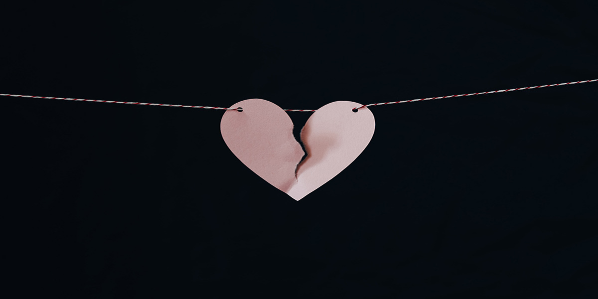 A pink paper heart, torn down the middle hangs from a thread against a jet black background